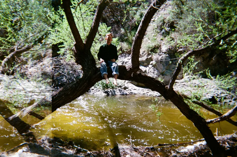 James Spaite sitting on a low tree branch above a green creek pool, trees and rocks all around.