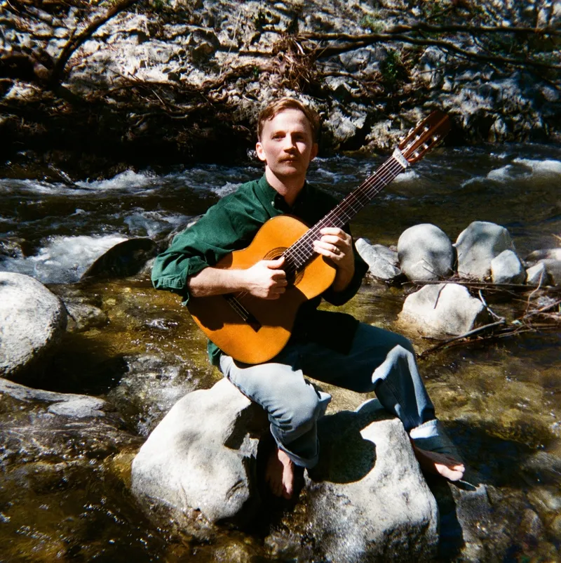 James Spaite sitting on a boulder in a creek, barefoot, a classical guitar across his lap, looking straight at the camera.