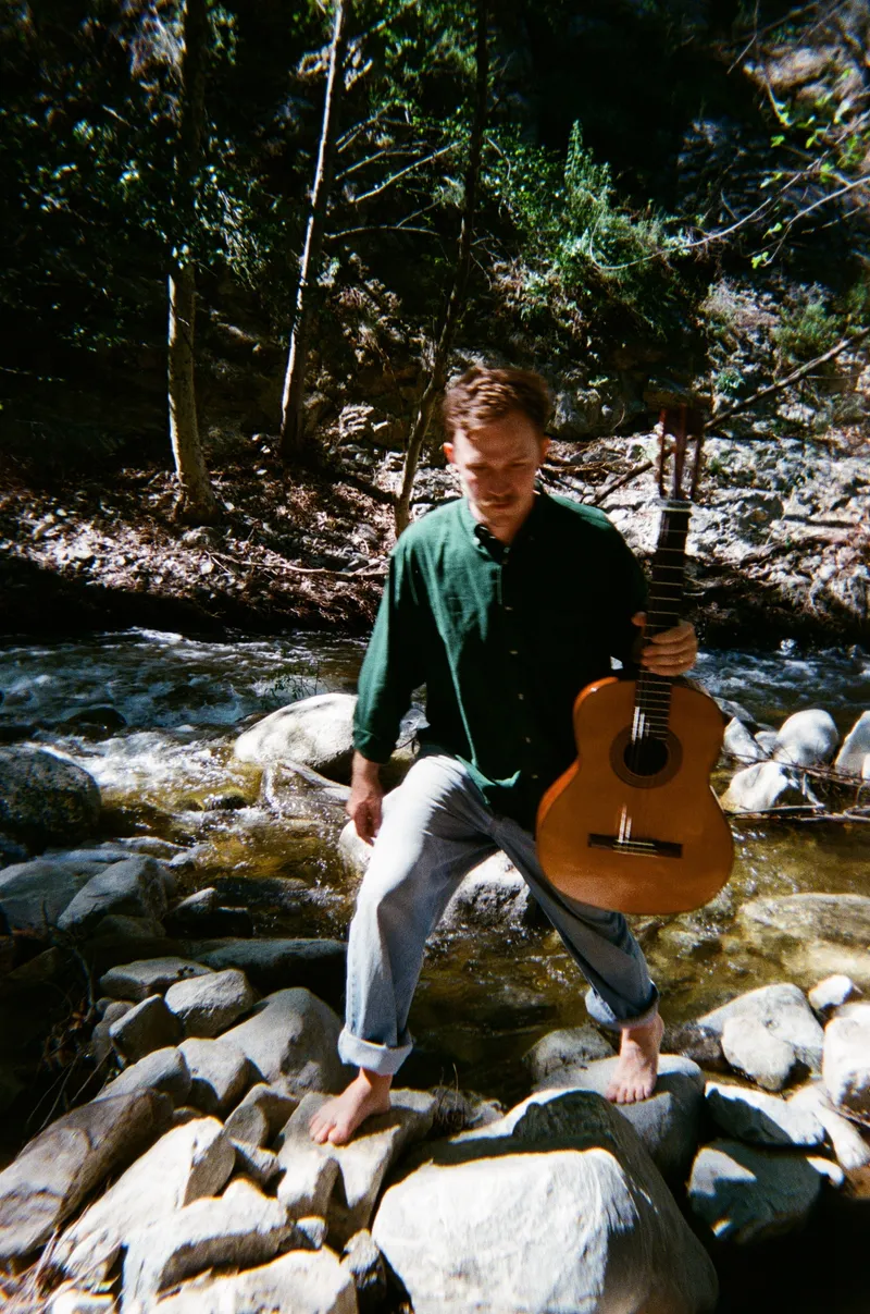 James Spaite standing barefoot on creek boulders, holding a classical guitar at his side.